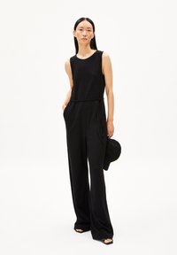 Schwarzer ärmelloser Jumpsuit mit taillierter Taille, fließenden, weiten Beinen und seitlichen Taschen. Das Model hält eine schwarze Kappe und steht vor einem weißen Hintergrund.