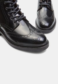 Botas de cuero negro con cordones que presentan un diseño brogue, un acabado suave, punta redonda y cordones texturizados con sutiles acentos perforados.