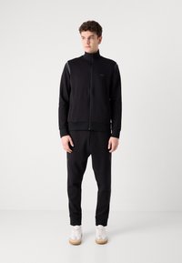 Jachetă neagră cu fermoar, guler înalt și manșete cu bordură, asociată cu pantaloni jogger asortați. Prezintă un logo subtil și adidași albi.