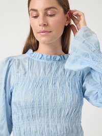 Γυναίκα με ανοιχτό καστανά μαλλιά, φοράει ένα ανοιχτό μπλε μακρυμάνικο πουκάμισο με ραφές, διορθώνει τα μαλλιά της κοντά στο δεξί αυτί της, όπου έχει ασημένια σκουλαρίκια.