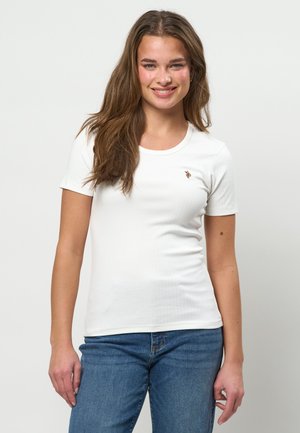 Ung kvinde med langt brunt hår, der smiler, iført en tætsiddende hvid kortærmet skjorte og blå jeans, stående foran en ensfarvet hvid baggrund.