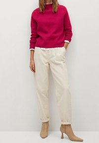 Fuchsia Pullover mit gerippter Textur, cremefarbene, lässig geschnittene Hose und beigefarbene, spitze Ankle Boots. Ein weißes Hemd blitzt am unteren Rand des Pullovers hervor.