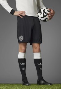 Black soccer shorts with ribbed texture and logo, paired with black and white striped knee-high socks, and black cleats, holding a soccer ball.