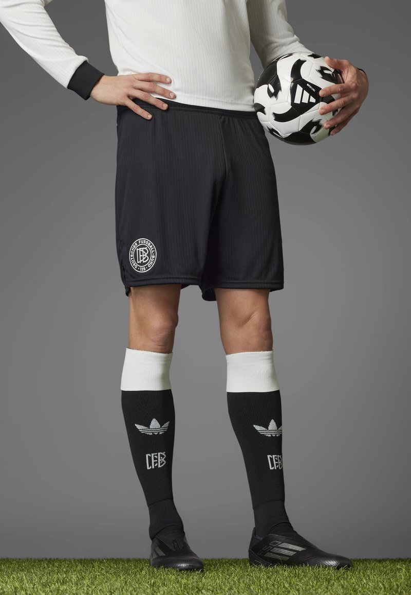 Black soccer shorts with ribbed texture and logo, paired with black and white striped knee-high socks, and black cleats, holding a soccer ball.