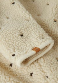 Tissu en polaire de couleur crème avec de petits motifs en forme de cœur noir et un bord roulé, comportant une petite étiquette en cuir marron.