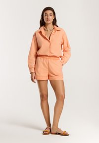 Completo di camicia e pantaloni corti color pesca in tessuto leggero. La camicia ha un colletto e chiusura a bottoni; i pantaloni corti hanno una vita elastica. Sandali con fasce decorative.