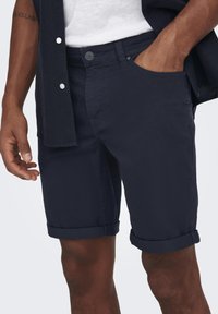 Marineblå shorts med oppbrettede ermer, laget av et lett stoff. Har knappelukking og frontlommer. Enkel, moderne design.