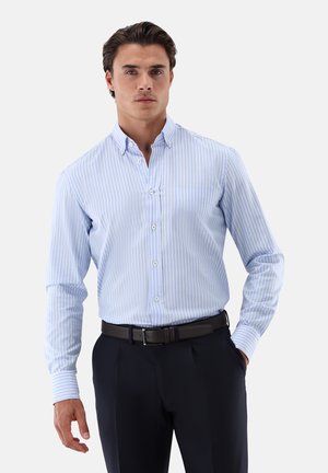Camicia a righe blu chiaro con colletto button-down, maniche lunghe e taschino, abbinata a pantaloni scuri e una cintura nera.
