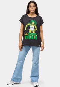 Camiseta negra con un gráfico de She-Hulk en verde y blanco, con texto amarillo. Combinada con pantalones vaqueros acampanados de color azul claro y zapatillas blancas.