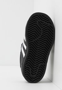 Mustat lenkkarit valkoisilla raidoilla; tasainen kumipohja, jossa on sahalaitakuvio; sivuprofiilissa näkyy teksturoitu materiaali ja ompeluyksityiskohdat.