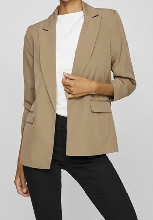Vrouw met een beige blazer met klepzakken over een wit shirt en zwarte broek, hand houdt het revers van de blazer vast.