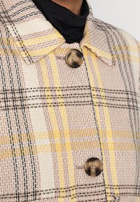 Kraagloze geblokte jas in beige, geel en zwart met geweven textuur en grote bruine knoopdetails.