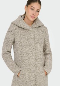 Femme portant un manteau en laine beige texturé avec un large col châle, les mains dans les poches, et des boucles d'oreilles en or.