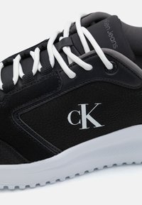 Crne tenisice s gornjim dijelom od teksturirane kože i antilopa, bijelim vezicama, potpisanim "ck" logom i bijelim gumenim potplatom s uzorkom.