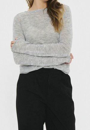 Femme portant un pull à manches longues gris clair et un pantalon noir ample, les bras croisés, sur un fond blanc uni.