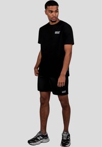 Černé tričko s malým bílým logem, odpovídající černé šortky a šedé tenisky. Jednoduchý design, sportovní střih, látka vypadá lehce.