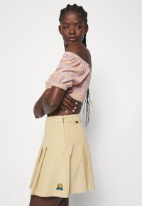 Λεβάντα τοπ με ανοιχτούς ώμους και κίτρινο σχέδιο, συνδυασμένο με μπεζ φούστα από κορντούρα με πιέτες και κεντητό λογότυπο βάτραχου στο τελείωμα.