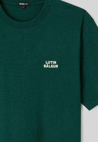 T-shirt vert foncé à manches courtes et col rond, avec un petit texte blanc "LUTIN RÂLEUR" sur le côté gauche de la poitrine, étiquette taille L à l'intérieur du col.