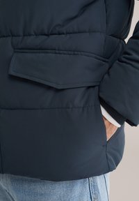 Marinblå vadderad jacka med en strukturerad yta. Har en sidoficka, ribbade muddar och en synlig fåll, kombinerad med ljusblå jeans.