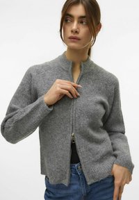 Cárdigan gris con cremallera y cuello acanalado en tejido texturizado. Presenta mangas largas y un dobladillo corto, combinado con jeans azules.