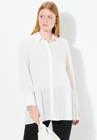Blusa bianca a maniche lunghe con chiusura a bottoni sul davanti, texture liscia e spacchi laterali, abbinata a pantaloni neri e una borsa di colore chiaro.