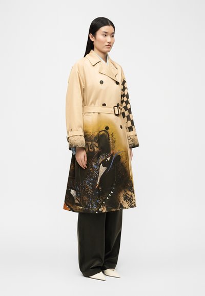 Γυναίκα που φοράει μπεζ παλτό βροχής με μαύρα κουμπιά, ζώνη, μοτίβο σκακιέρας σε ένα μανίκι και καλλιτεχνικό σχέδιο πουλιού στο κάτω μέρος.
