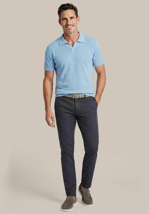 Man staat, draagt een lichtblauw polo met korte mouwen, donkere broek met riem en grijze instapschoenen, glimlachend tegen een effen beige achtergrond.