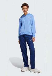 Gaiši zils ar rāvējslēdzēju krekls ar priekšējo kabatu un baltu logo uz krūtīm, apvienots ar tumši ziliem sporta biksēm un baltām sporta apaviem.