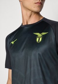Mann iført svart sportstrøye med neon gul S.S. Lazio ørn-logo og Mizuno-emblem på brystet, vender seg litt til siden.