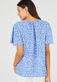 Blauwe bloemen blouse met korte mouwen, een gerimpelde achterkant bij de hals en witte bloemenpatronen. De blouse heeft een ontspannen pasvorm en een zachte stoftextuur.