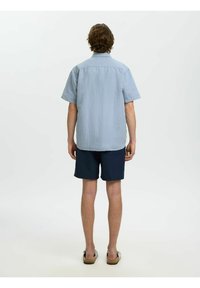 Homme aux cheveux bruns portant une chemise à manches courtes rayée bleu clair, un short bleu marine et des sandales, debout de dos sur un fond blanc uni.