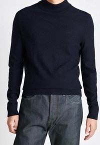 Mörkblå polotröja i mjukt stickat material med ribbade ärmslut och nederkant; har en subtil logotyp på bröstet; kombinerad med mörka jeans.