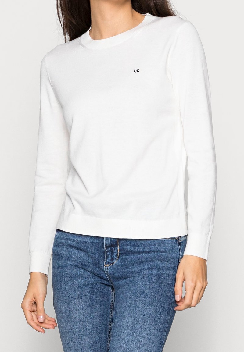 Pull en tricot blanc à manches longues avec un col rond et des poignets côtelés. Présente un petit logo "CK" noir sur la poitrine. Porté avec un jean bleu.