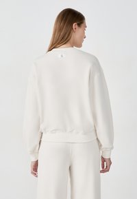 Sudadera de color beige claro con un ajuste holgado, puños y dobladillo acanalados. Presenta una pequeña etiqueta en la parte posterior del cuello. Textura de tela suave y lisa.