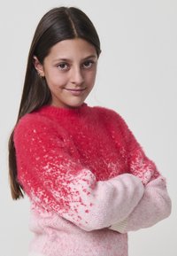 Jeune fille aux longs cheveux bruns et petites boucles d'oreilles en or, portant un pull en laine rouge et rose, souriante avec les bras croisés.