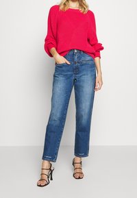 Femme portant un pull rose vif, un jean bleu à jambes droites avec une main dans la poche, et des sandales à talons hauts noires à brides, sur fond blanc.