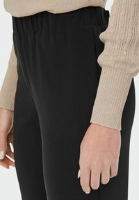 Zwarte rekbare legging met een elastische tailleband, gecombineerd met een beige gebreide trui met textuur. Close-up van de details van de outfit.