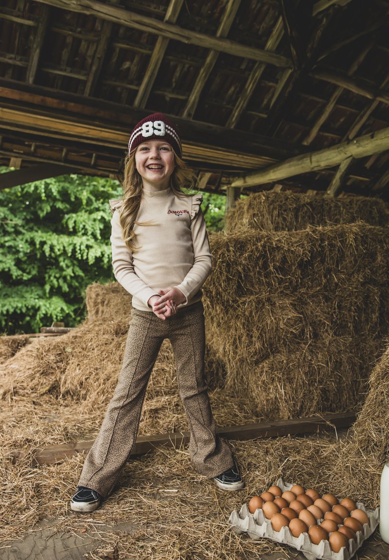 Kind met een beige, gerimpeld T-shirt met lange mouwen met de tekst "Amour", gecombineerd met bruine gemusterde flared broeken en zwarte sneakers, staand in het hooi.