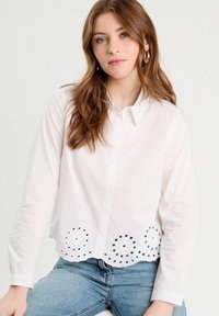 Chemise blanche à manches longues, dotée de découpes décoratives en œillets le long de l'ourlet et d'un col.