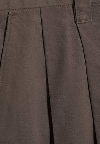 Gros plan d’un tissu plissé marron avec des coutures visibles et une boucle de ceinture, montrant la texture et la structure du matériau textile.