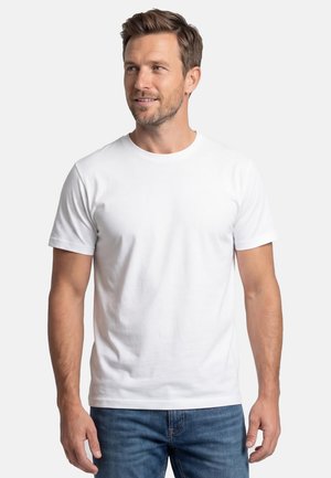 Man met kort bruin haar, die een effen witte T-shirt met korte mouwen en een blauwe spijkerbroek draagt, kijkt iets naar rechts, neutrale achtergrond.