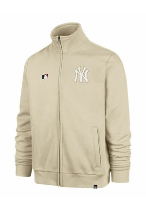 Chaqueta beige con cremallera hecha de un tejido suave que presenta un cuello alto. En la parte delantera muestra un logo blanco del equipo y el emblema de la MLB. Incluye bolsillos laterales.