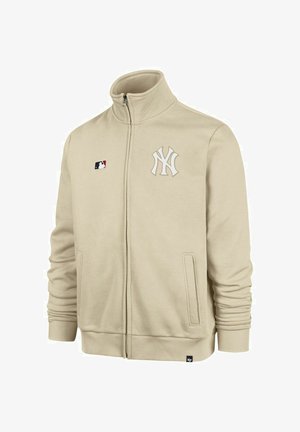 Giacca beige con zip realizzata in un tessuto morbido, presenta un colletto alto. Sul davanti è visibile un logo bianco della squadra e l'emblema MLB. Include tasche laterali.