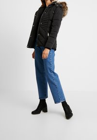 Giubbotto puffer nero con cappuccio, caratterizzato da un bordo in pelliccia sintetica, trapuntatura a zig-zag e accessori in metallo argentato. Abbinato a jeans blu e stivaletti neri.