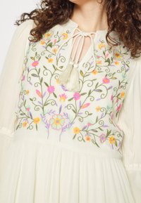 Broderad krämfärgad klänning med blommönster i livliga rosa, gula och gröna trådar, knytning i halsringningen och flödande textil med struktur.