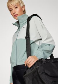 Chaqueta ligera con cremallera en verde menta y gris, con puños elásticos, llevada con un bolso de viaje negro, que presenta detalles de malla.