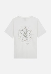 Witte katoenen t-shirt met een zwart en gouden bloemenontwerp op de achterkant, met geometrische lijnen en een handtekening onderaan.
