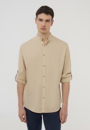 Giovane uomo con capelli corti e scuri che indossa una camicia beige con bottoni, maniche arrotolate e pantaloni scuri su uno sfondo chiaro e uniforme.