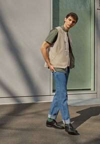 Beige gewatteerde vest over een groen T-shirt met korte mouwen, gecombineerd met opgerolde blauwe jeans en zwarte schoenen. Minimalistisch ontwerp met een gladde textuur.