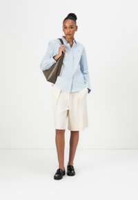 Lyseblå skjorte med knapper, kremfargede shortser, svarte loafers og olivengrønn toteveske. Glatt stoff og ledig passform, med subtile detaljer.